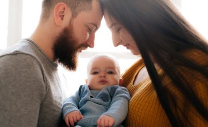 a man with a beard and a woman with long dark hair look down at a baby wearing blue between them